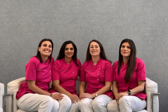 Quattro donne, Assistenti Coppedè Medical Hub di Roma, indossano top rosa acceso e pantaloni bianchi abbinati, sedute su un divano bianco su uno sfondo grigio, sorridono e posano per una foto di gruppo.