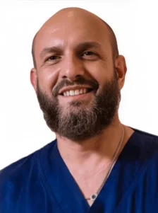Un uomo sorridente con la testa calva e la barba folta, che indossa un camice da medico blu navy e una collana, è raffigurato su uno sfondo bianco semplice presso il Polo Medico Dott. Emiliano De Sanctis Coppedè di Roma.