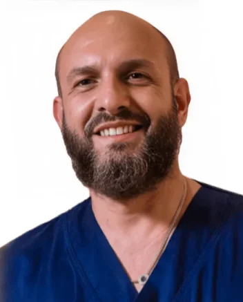 Un uomo sorridente con la testa calva e la barba folta, che indossa un camice da medico blu navy e una collana, è raffigurato su uno sfondo bianco semplice presso il Polo Medico Dott. Emiliano De Sanctis Coppedè di Roma.