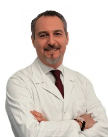 Il Dott. Edoardo Brauner del Coppedè Medical Hub di Roma, un uomo con capelli corti e scuri e pizzetto grigio, sorride in camice bianco e cravatta rossa, incrociando le braccia su uno sfondo bianco.