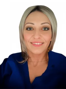 La Dott.ssa Flavia Aghilone Coppedè sorride all'obiettivo, con i suoi capelli biondi lunghi fino alle spalle e gli occhi azzurri completati da un top blu e una delicata collana, su uno sfondo bianco semplice al Medical Hub di Roma.