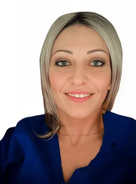 La Dott.ssa Flavia Aghilone Coppedè sorride all'obiettivo, con i suoi capelli biondi lunghi fino alle spalle e gli occhi azzurri completati da un top blu e una delicata collana, su uno sfondo bianco semplice al Medical Hub di Roma.