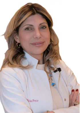 La Dott.ssa Valentina Prece del Coppedè Medical Hub di Roma, una donna dai capelli biondi lunghi fino alle spalle con un camice bianco, si erge sicura di sé con le braccia incrociate e un caldo sorriso su un semplice sfondo bianco.
