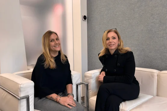 Due donne siedono su moderne sedie bianche in una stanza minimalista dell'Amministrazione Coppedè Medical Hub di Roma. Entrambe hanno lunghi capelli biondi e sorridono all'obiettivo: una indossa un top nero, l'altra una giacca nera e pantaloni scuri.