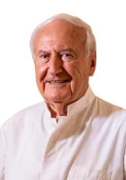 Il Dott. Arnold Brauner, un uomo anziano con i capelli bianchi, è sorridente e indossa una camicia bianca, in piedi davanti a uno sfondo bianco.