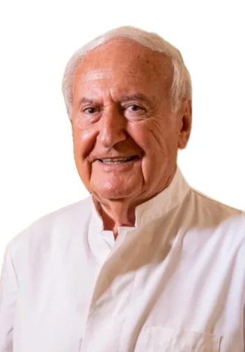 Il Dott. Arnold Brauner, un uomo anziano con i capelli bianchi, è sorridente e indossa una camicia bianca, in piedi davanti a uno sfondo bianco.