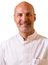 Un uomo sorridente, il Dott. Carlo Rasiel Brauner del Coppedè Medical Hub di Roma, indossa un camice da cuoco bianco abbottonato con una penna nel taschino, su uno sfondo bianco.
