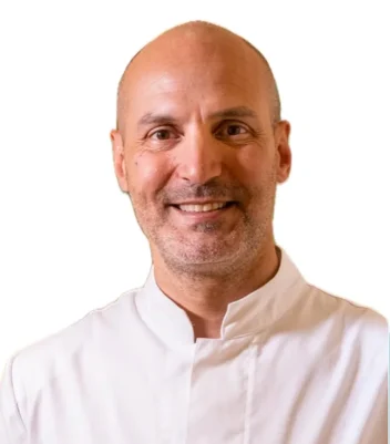 Un uomo sorridente, il Dott. Carlo Rasiel Brauner del Coppedè Medical Hub di Roma, indossa un camice da cuoco bianco abbottonato con una penna nel taschino, su uno sfondo bianco.