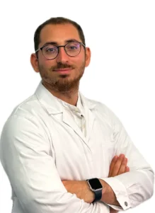 Il Dott. Claudio Saulli, un uomo con capelli corti, barba e occhiali, indossa un camice bianco e incrocia le braccia, in piedi su un semplice sfondo bianco.