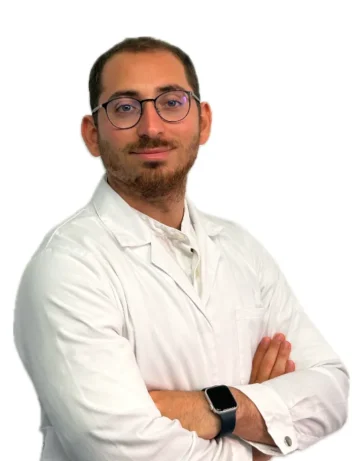 Il Dott. Claudio Saulli, un uomo con capelli corti, barba e occhiali, indossa un camice bianco e incrocia le braccia, in piedi su un semplice sfondo bianco.