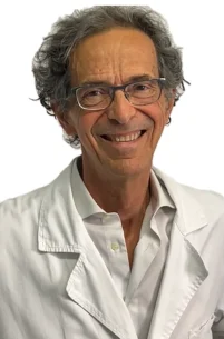 Il Dott. Guido Donati del Coppedè Medical Hub di Roma, un uomo anziano con capelli grigi e ricci e occhiali, sorride indossando un camice bianco e una camicia, in piedi contro uno sfondo bianco semplice.