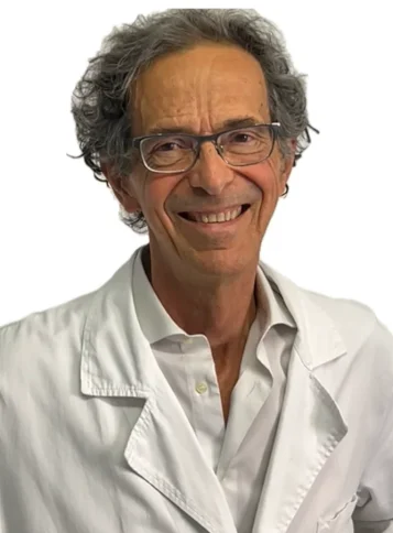 Il Dott. Guido Donati del Coppedè Medical Hub di Roma, un uomo anziano con capelli grigi e ricci e occhiali, sorride indossando un camice bianco e una camicia, in piedi contro uno sfondo bianco semplice.