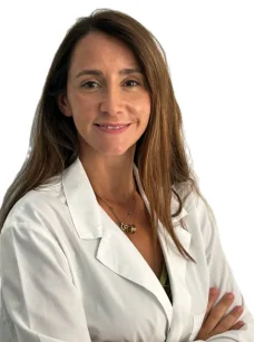 La Dott.ssa Claudia Milella Coppede, del Medical Hub Roma, con lunghi capelli castani e camice bianco, sta in piedi con le braccia incrociate e sorride alla telecamera su uno sfondo bianco.