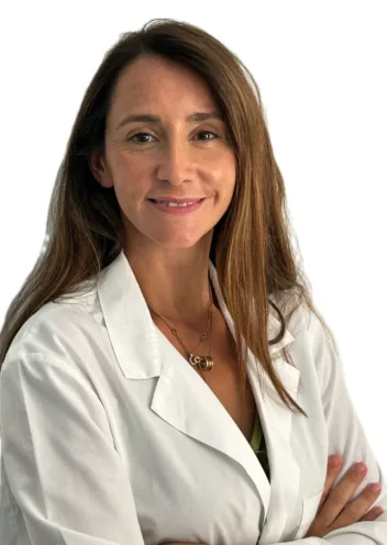 La Dott.ssa Claudia Milella Coppede, del Medical Hub Roma, con lunghi capelli castani e camice bianco, sta in piedi con le braccia incrociate e sorride alla telecamera su uno sfondo bianco.