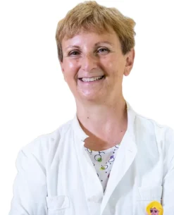 La Prof.ssa Maria Teresa Fadda, una persona sorridente con i capelli corti, indossa un camice bianco e una camicia a fantasia, in posa su uno sfondo bianco semplice.