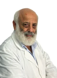 Uomo anziano con testa calva e barba bianca che indossa un camice bianco e una camicia azzurra, in posa su un semplice sfondo bianco presso il Centro Medico Dott. Guido Coen Coppede di Roma.