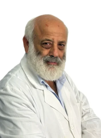 Uomo anziano con testa calva e barba bianca che indossa un camice bianco e una camicia azzurra, in posa su un semplice sfondo bianco presso il Centro Medico Dott. Guido Coen Coppede di Roma.